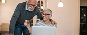 Senior woman sits at laptop while senior man looks over her shoulder.