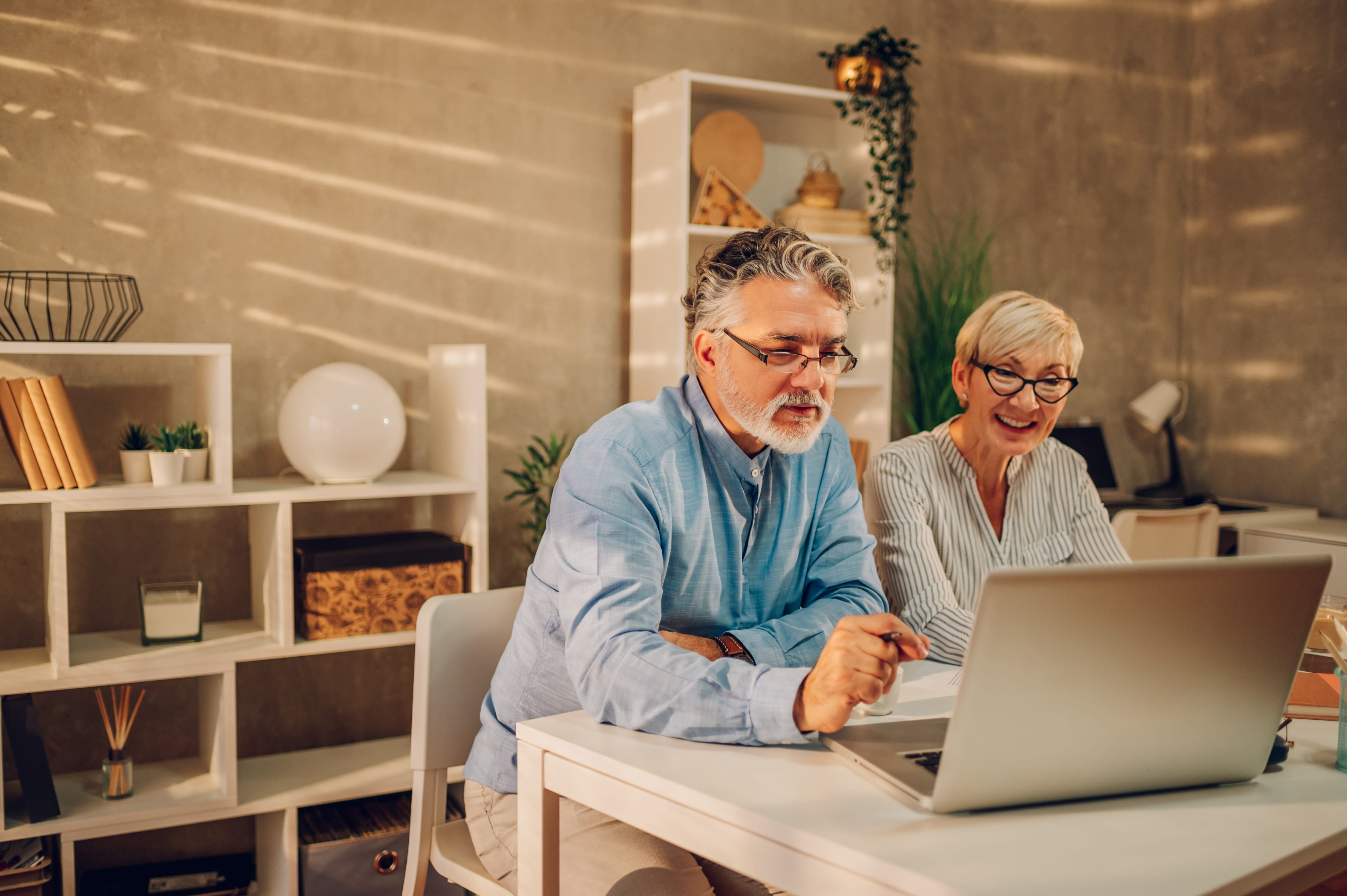 Two seniors sit at a laptop in a home office.