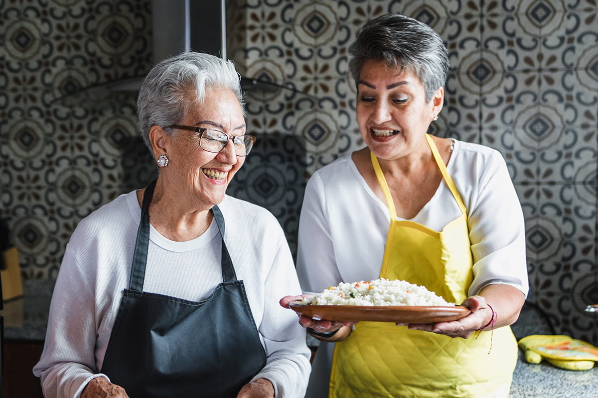 Two senior women wearing aprons look at a dish of food.